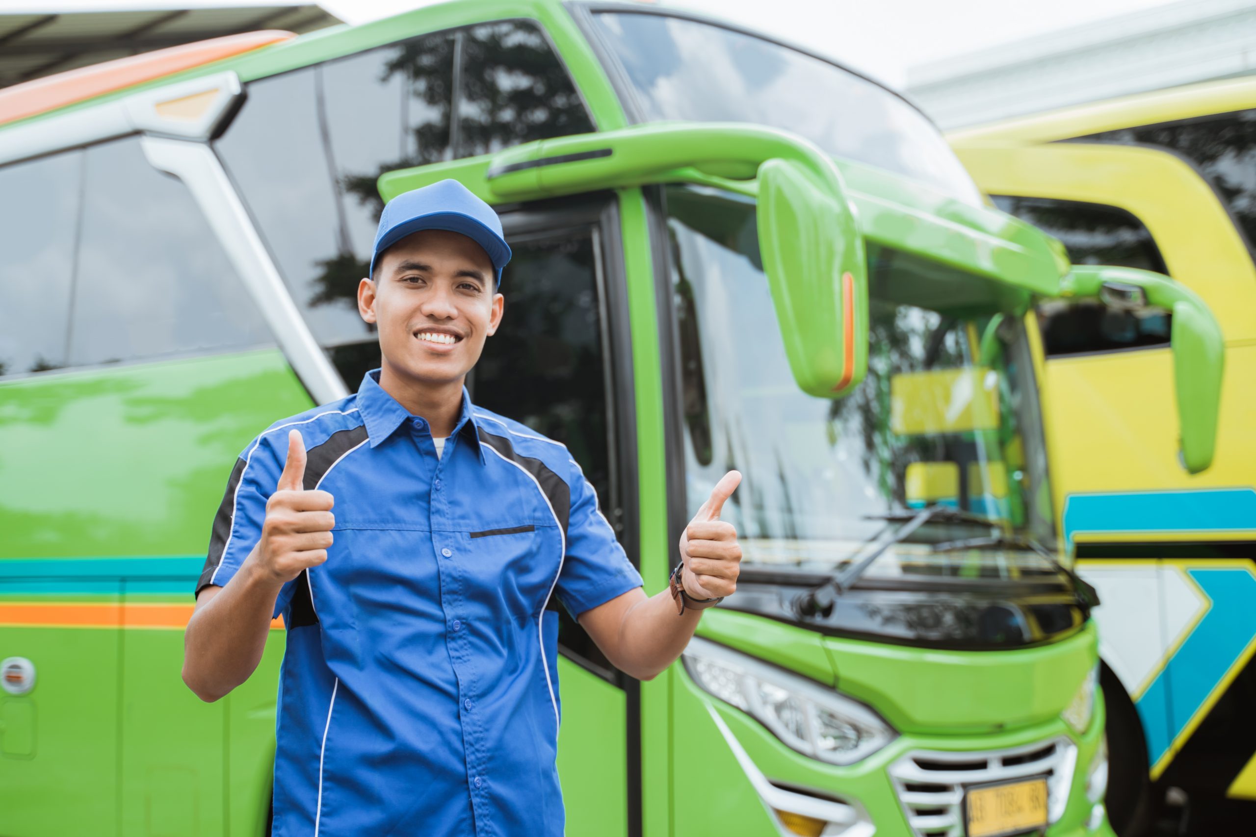 A handsome bus driver in uniform and hat smiles with thumbs up against the background of the bus